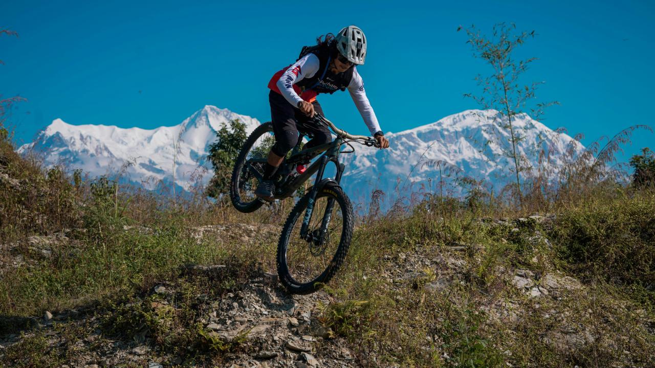 Mountain biker jumps over a dirt mound with snow-capped mountains.